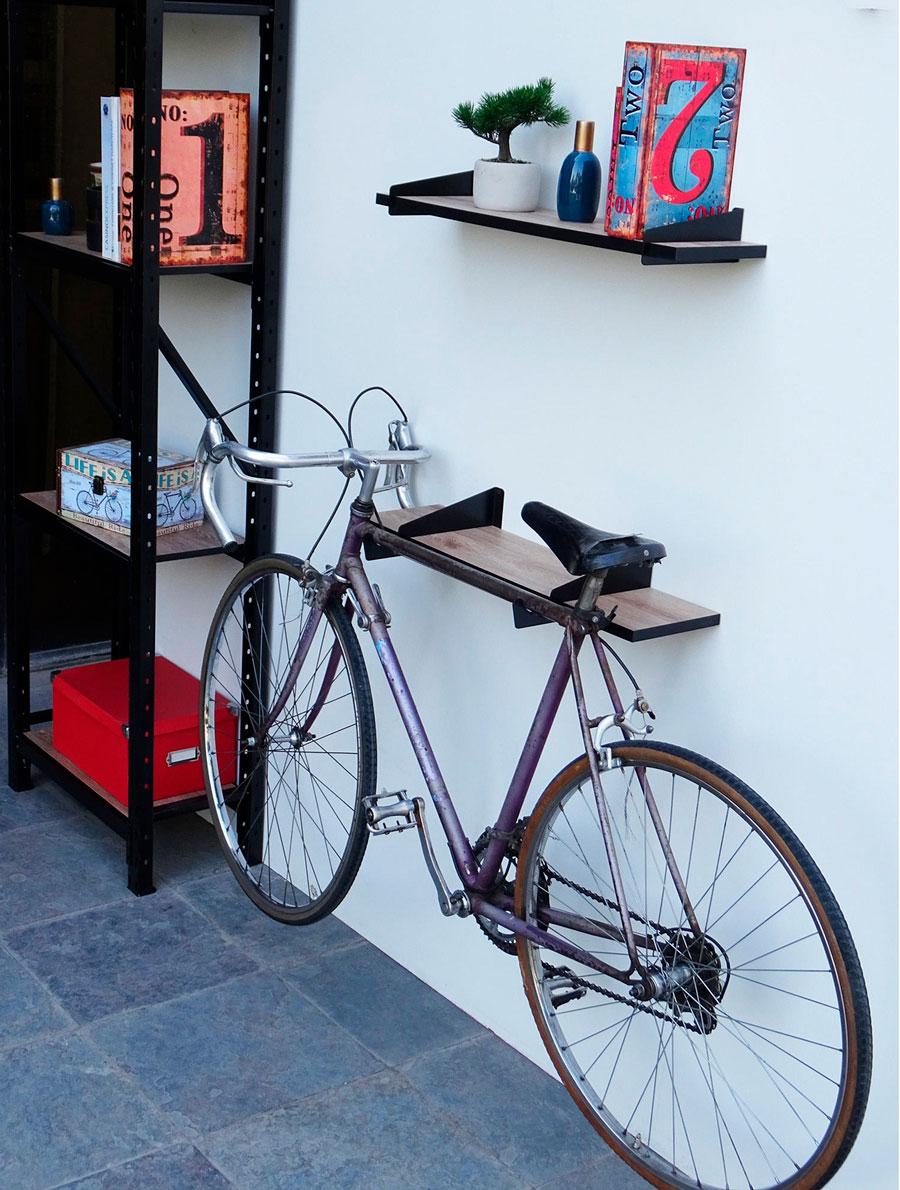 Muro con dos estantes de madera y marcos de metal negro. En una hay un libro y una maceta y en el segundo está enganchado una bicicleta.