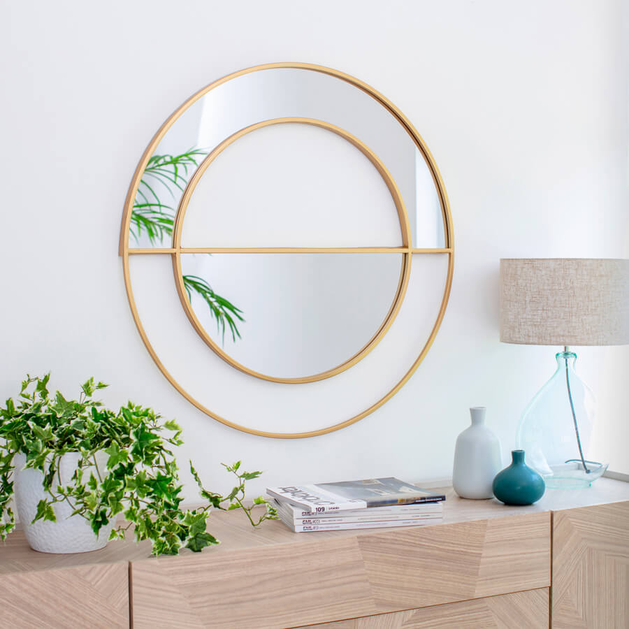 Recibidor con paredes blancas y un mueble de madera con revistas, planta, jarrones una lámpara. Sobre el mueble hay un gran espejo redondo con marco y detalles dorados.