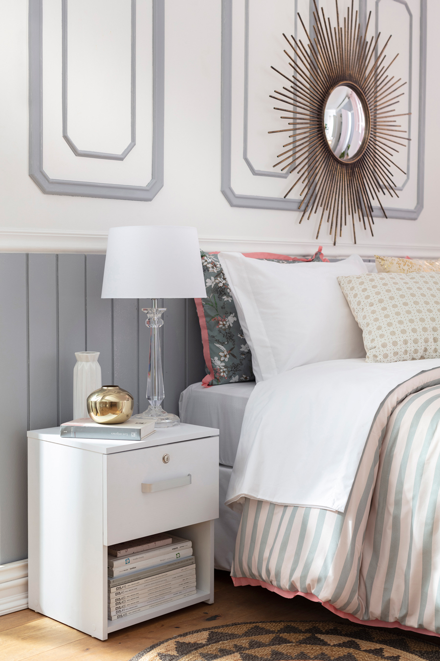 Detalle de un dormitorio con una pared con molduras blancas y grises. Hay un velador blanco junto a la cama, la que tiene cojines y un plumón en tonos rosados y azul grisáceo. 