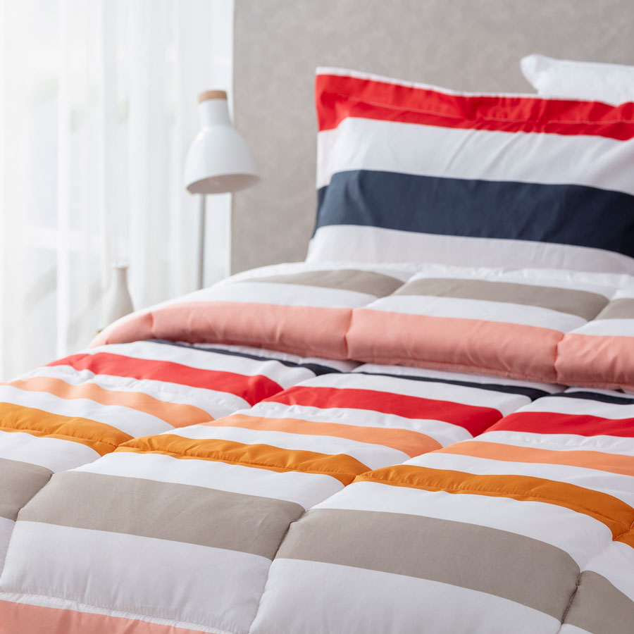 Cubre cama de líneas blancas, beige, anaranjado, salmón, rosado, rojo y azul marino. Las almohadas tienen el mismo diseño. 