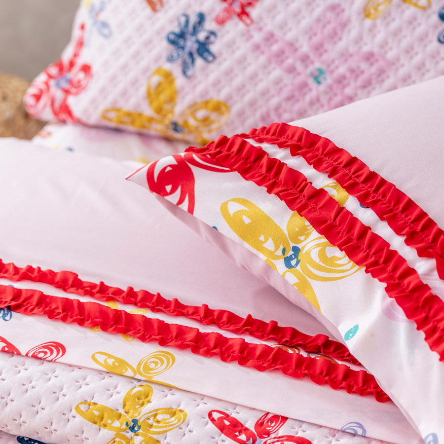 Detalle de una sábana blanca con flores rojas, azules y amarillas y un detalle rojo en las costuras. El quilt de esta cama tiene el mismo diseño de las sábanas, al igual que los cojines.