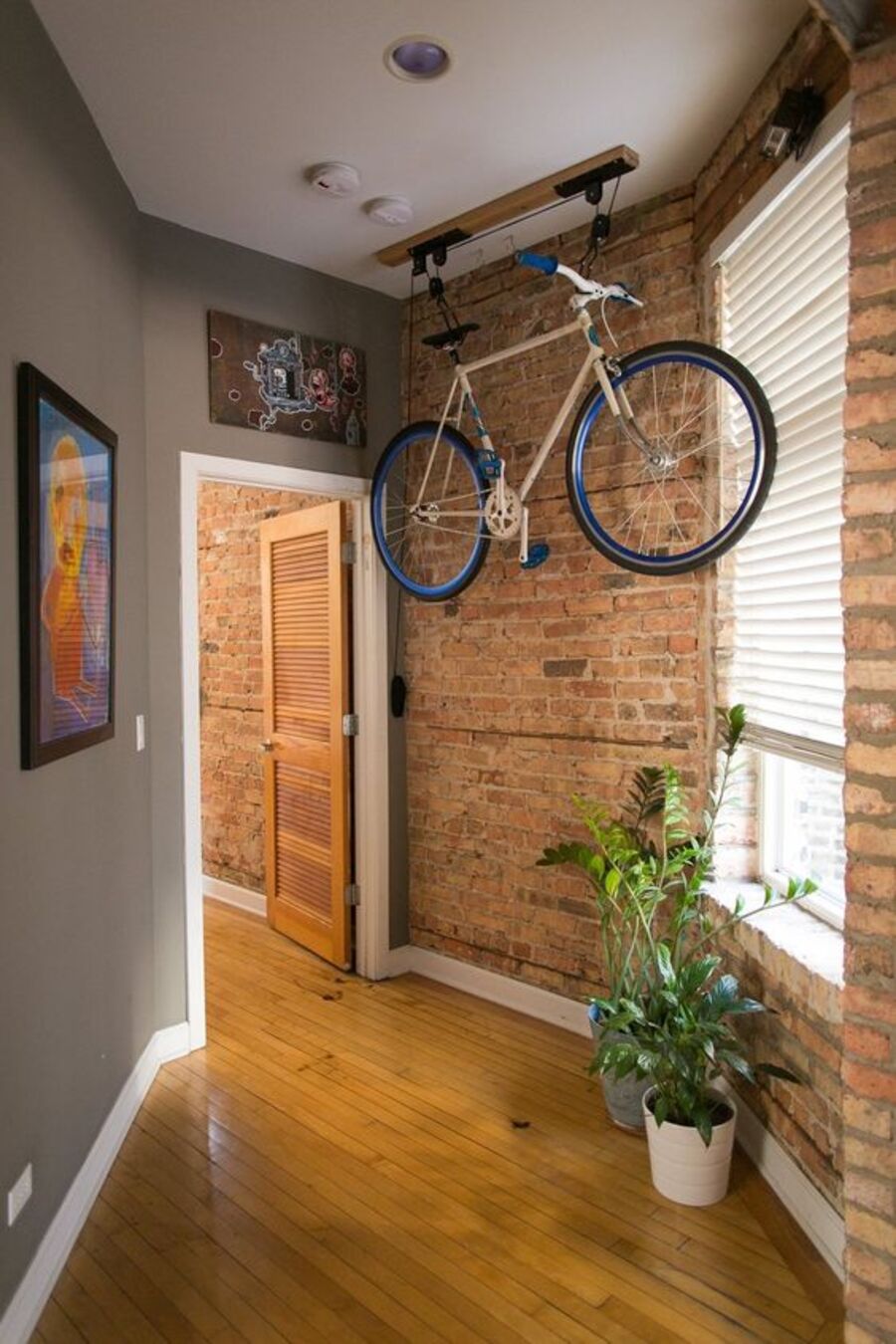Pasillo de piso de madera, pared izquierda gris, pared derecha de ladrillos y techo blanco. Desde este último cuelga una bicicleta desde una estructura con sistema de poleas. Hacia el fondo se ubica una puerta abierta de madera.