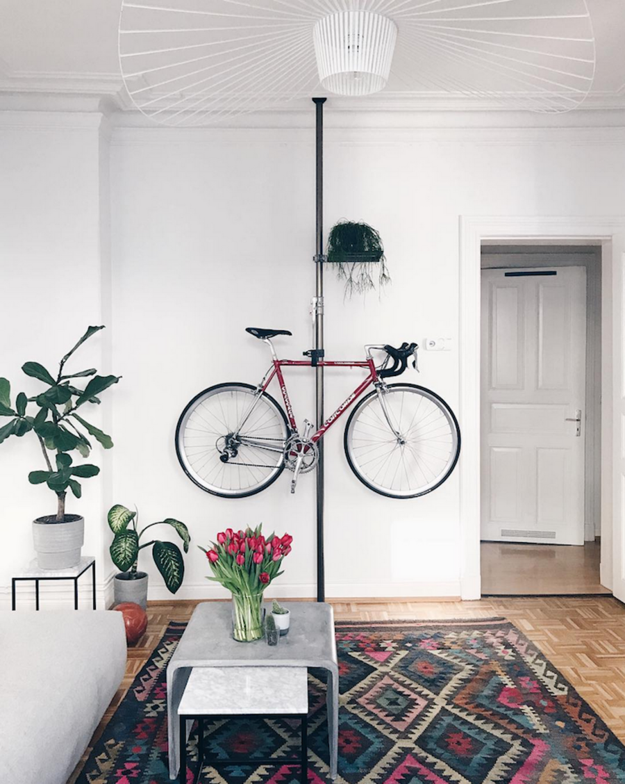 Sala de estar con paredes blancas, piso de madera, alfombra estampada de colores, sofá y mesa de centro gris claro, plantas decorativas y un tubo de fierro pegado a la pared del fondo. Desde este tubo cuelga una bicicleta roja. 