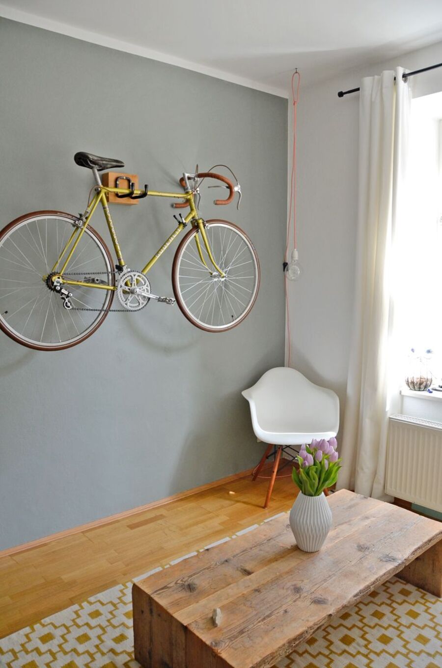 Rincón de una sala de estar con piso de madera, alfombra amarilla con blanco, mesa de centro café y silla plástica blanca. En una de sus paredes hay un perchero que sostiene una bicicleta pistera amarilla.
