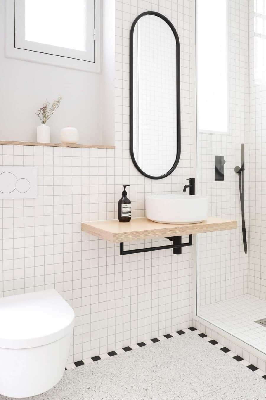 Baño con paredes de lozas blancas, suelo gris y un espejo ovalado grande con marcos negros.