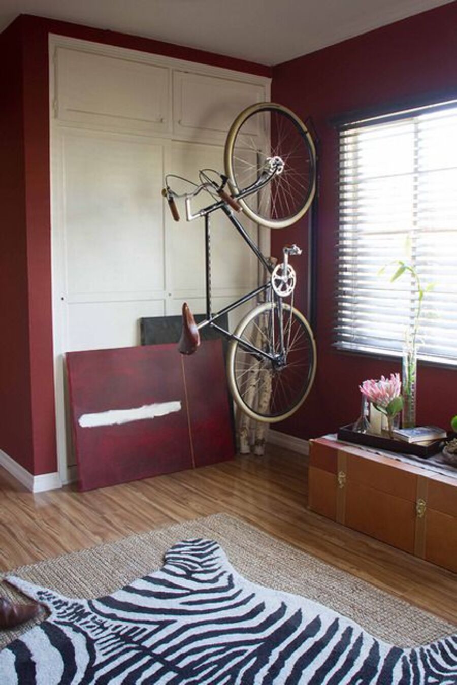 Rincón de una habitación con paredes burdeos, closet blanco empotrado, piso de madera, ventana con persianas y una bicicleta colgando verticalmente desde el espacio entre la ventana y el closet.
