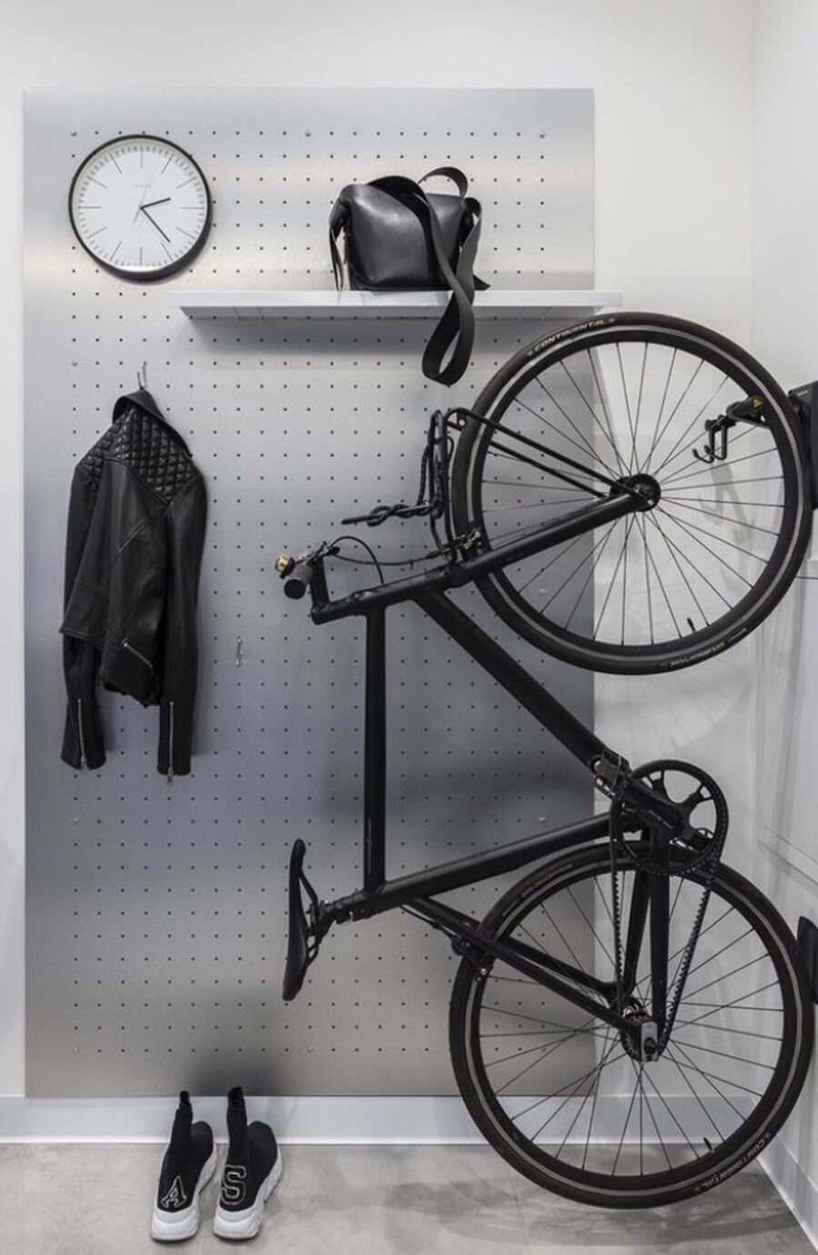 Bicicleta negra colgando desde una pared. Detrás de ella hay un recibidor con repisa, reloj y colgador.