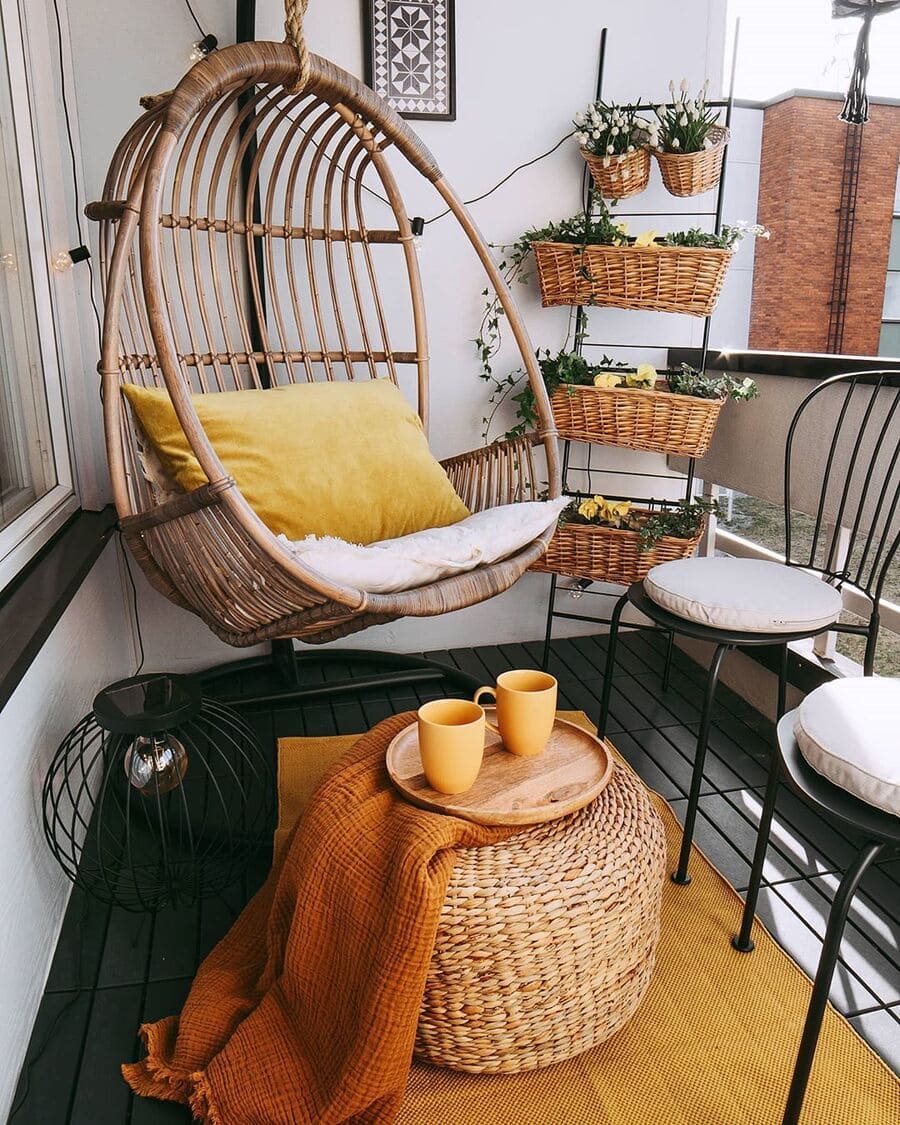 Balcón pequeño con una silla colgante de terraza con un cojín mostaza, hay una alfombra color mostaza, un pouf redondo e mimbre y sobre ella una manta naranja y una bandeja con dos tazas mostaza, como también dos sillas de metal negro.