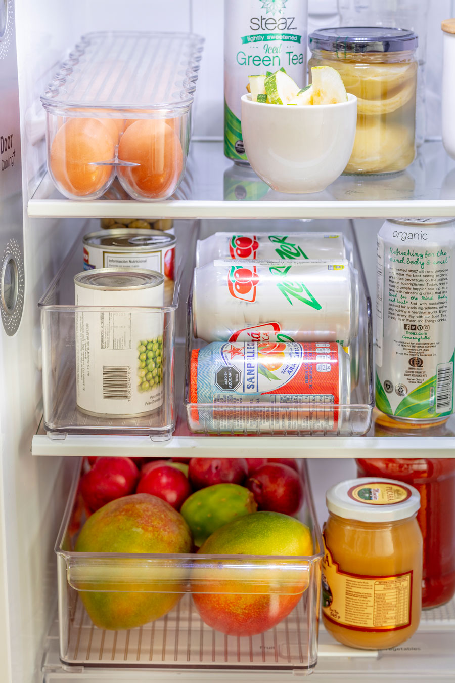 Cajas organizadoras plásticas transparentes dentro de un refrigerador. En su interior hay diferentes alimentos, tales como: huevos, latas de conserva, latas de bebidas y frutas.