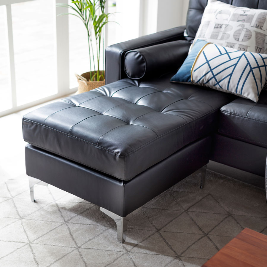 Detalle de un sofá en L de cuero negro. Está sobre una alfombra gris claro con líneas grises oscuras que forman triángulos. Sobre el sofá hay dos cojines con diferentes estampados, uno es de letras grises y el otro, blanco con líneas azules y un detalle en dorado.