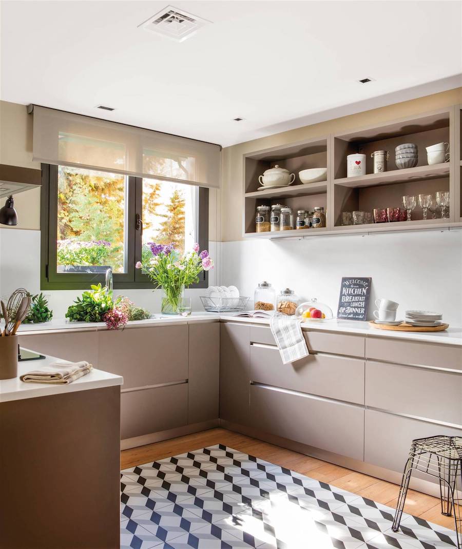 Cocina con muebles grises, piso de madera con una sección de piso vinílico con patrón blanco, negro y gris. La ventana de la cocina tiene marco negro y una cortina roller semi cerrada de color café claro. 