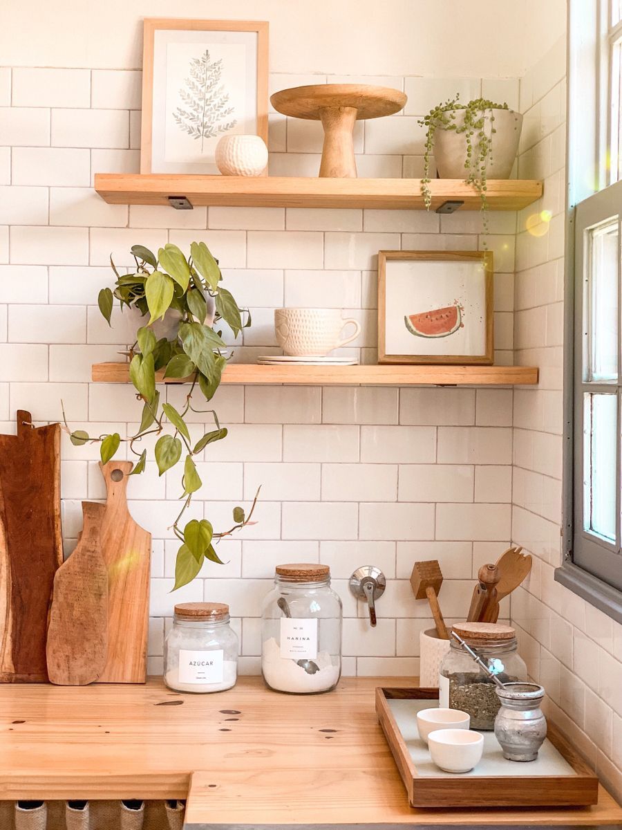 Detalle de una cocina con azulejos blancos, repisas de madera y encimera de madera. Sobre esta última hay tablas de madera, frascos y recipientes. 