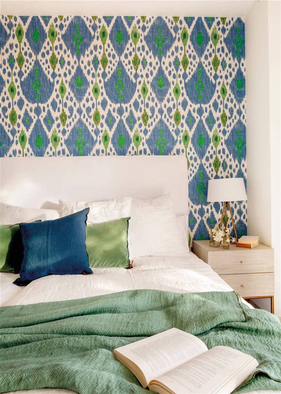 Dormitorio con cama de sábanas blancas, manta verde y cojines azules, que contrastan con un cojín azul petróleo. La pared del fondo tiene un papel con diseño verde y azul petróleo.