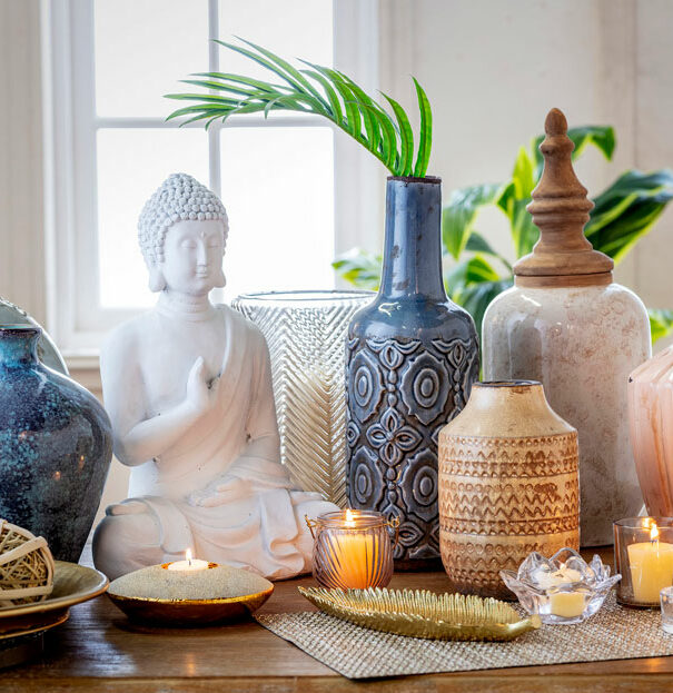 Detalle de la superficie de un mueble que sostiene frascos, velas y floreros en tonos neutros, así como una figura de Buda blanca.