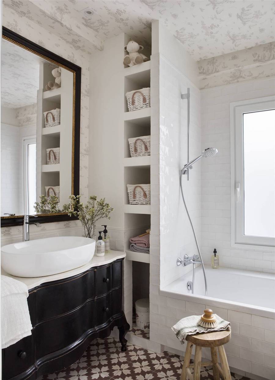 Baño de estilo clásico con tina blanca y azulejos blancos. El lavamanos es blanco y está sobre un mueble negro de estilo clásico. Junto a la tina hay un piso de madera con una toalla y una escobilla.
