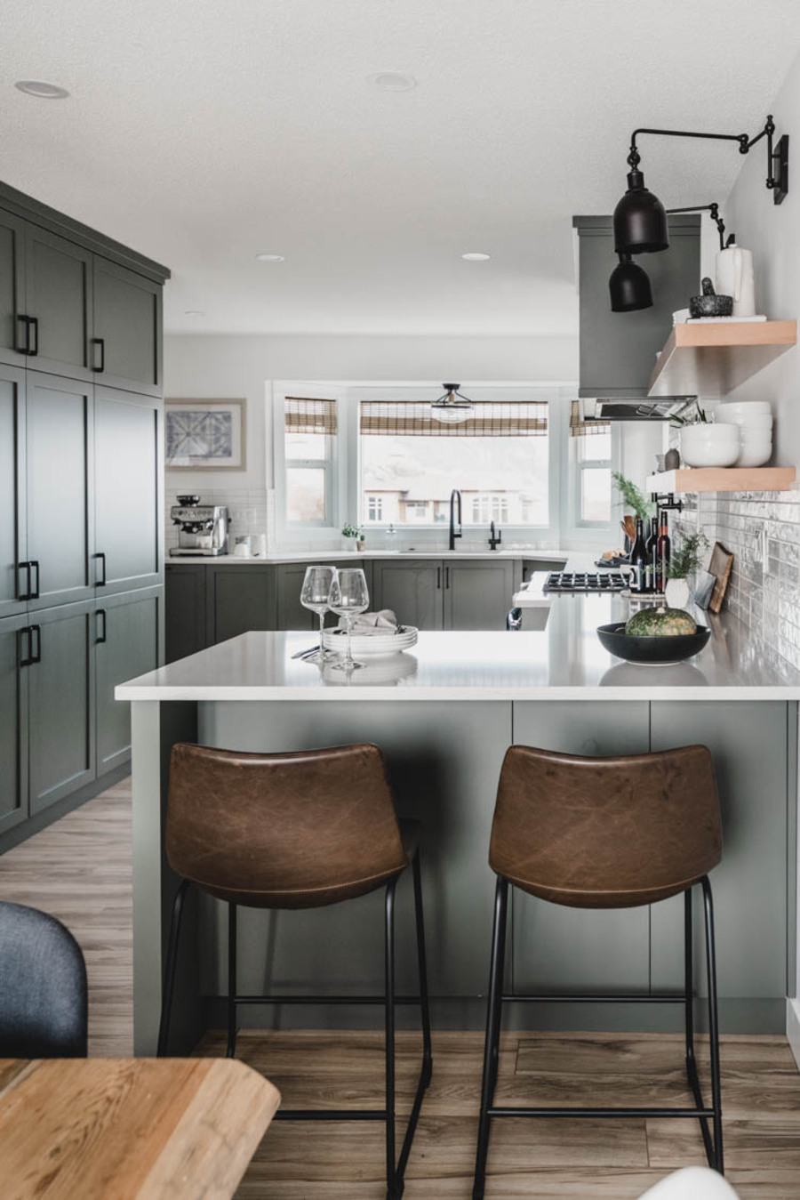 Cocina de concepto abierto con muebles de color verde grisáceo y encimera blanca. Los muros y azulejos son blancos y el piso, de madera.