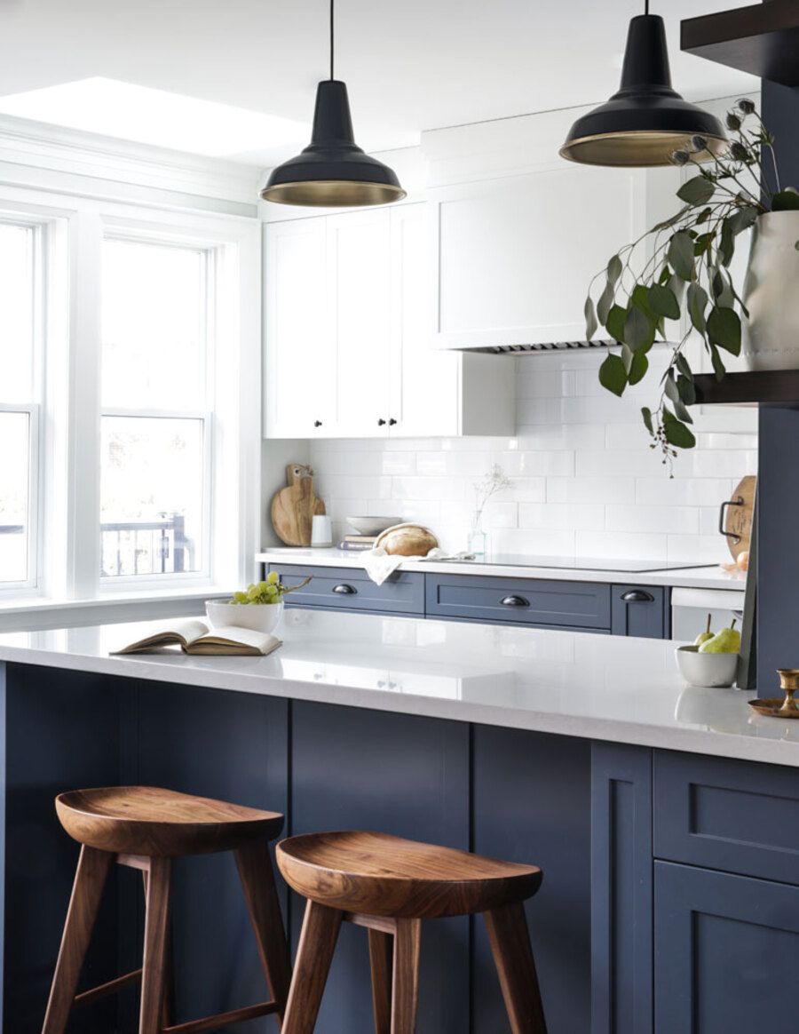 Cocina de concepto abierto con muebles inferiores azul marino, muebles superiores blancos y cubierta blanca. Desde el techo cuelgan dos lámparas metálicas negras de estilo industrial. 