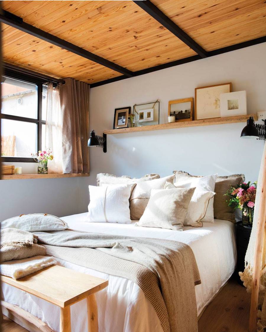 Dormitorio con techo y piso de madera, vigas negras y muro gris claro. La ropa de cama es de tonos neutros y sobre ella hay una repisa flotante de madera con cuadros y marcos de fotos.