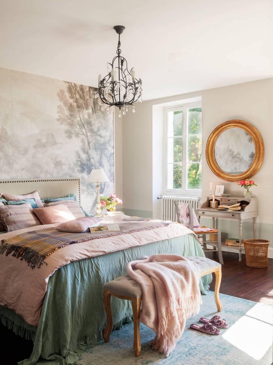 Dormitorio de estilo clásico con una cama con cobertor verde azulado, mantas y cojines rosados. Desde el techo cuelga una lámpara negra y junto a la cama hay un escritorio con un gran espejo dorado redondo sobre él. 