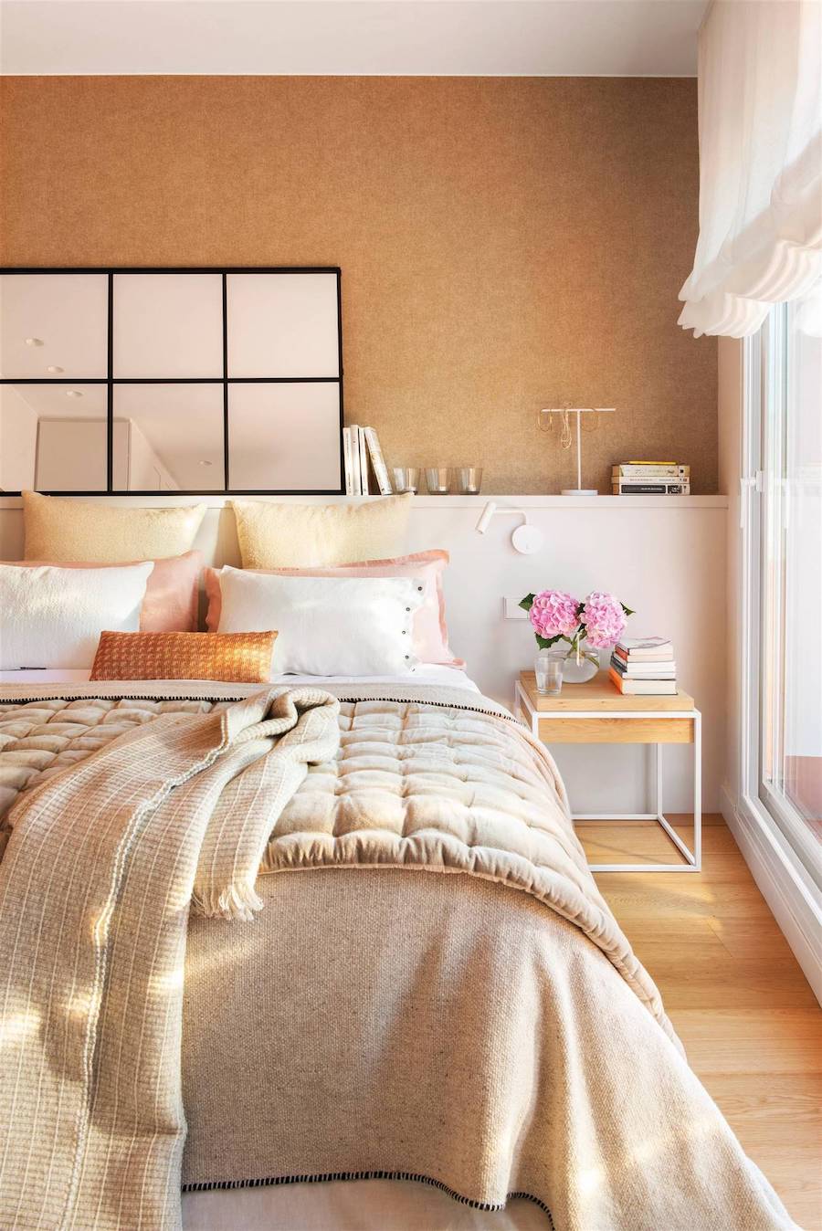 Dormitorio con ropa de cama entonos negros y arena, cojines amarillos, rosados, blancos y terracota. Sobre la cama hay un espejo tipo ventana con marco negro.