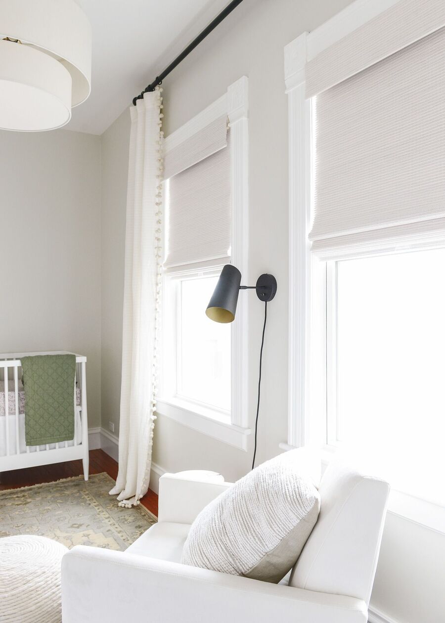 Ventanas de marco blanco con cortinas roller de color beige claro. Las paredes son del mismo color de las cortinas. Hay un sillón blanco con un cojín en tonos neutros, un pouf del mismo estilo del cojín, una alfombra y una cuna blanca.