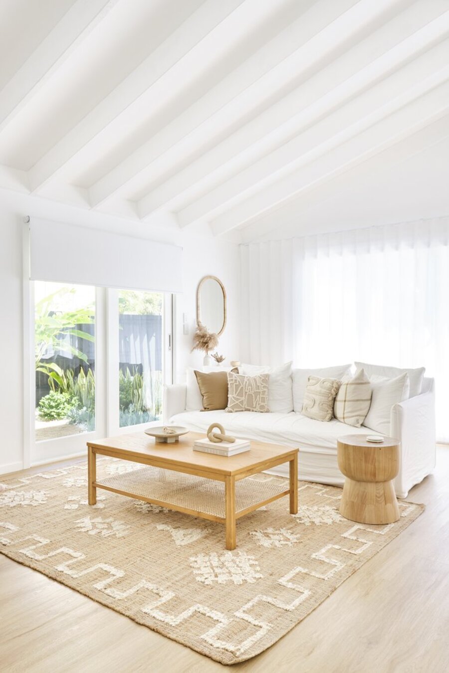 Sala de estar con techos, cortinas y muros blancos, piso de madera y una gran alfombra de yute con diseños blancos. Sobre ella hay una mesa de centro de madera clara y frente a ella, un sofá blanco con cojines en tonos tierra.