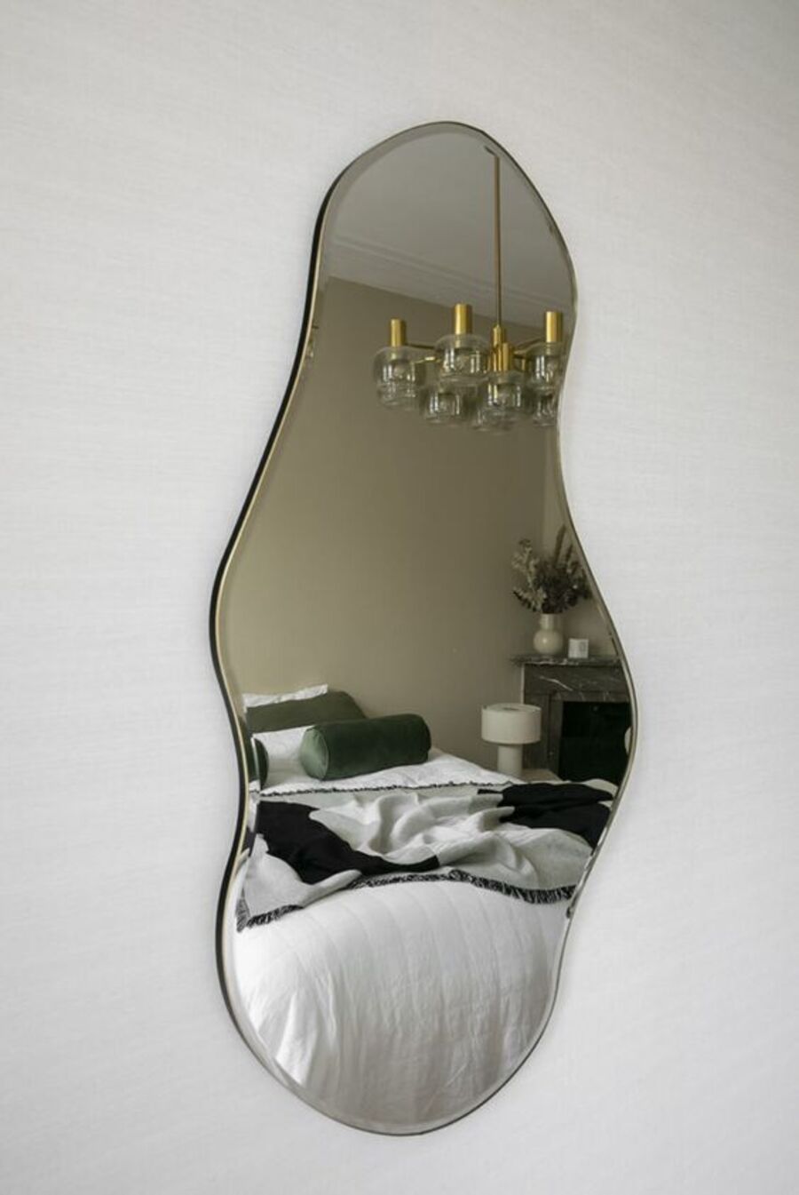 Pared blanca con un espejo con curvas que refleja una cama con cojines verdes y una lámpara colgante de estilo vintage.