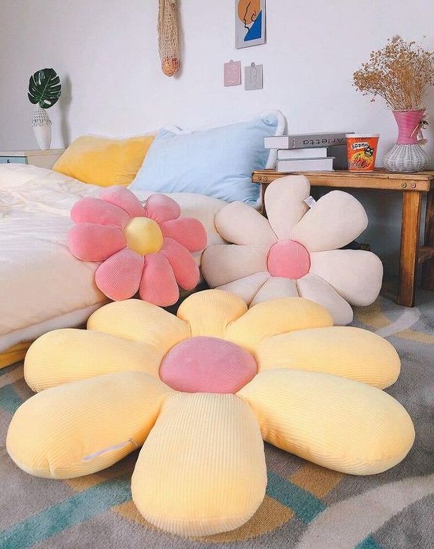 Tres cojines con forma de flor y en colores cálidos, como amarillo, rosado y beige. Están en el suelo y hacia el fondo hay una cama y un velador de madera.