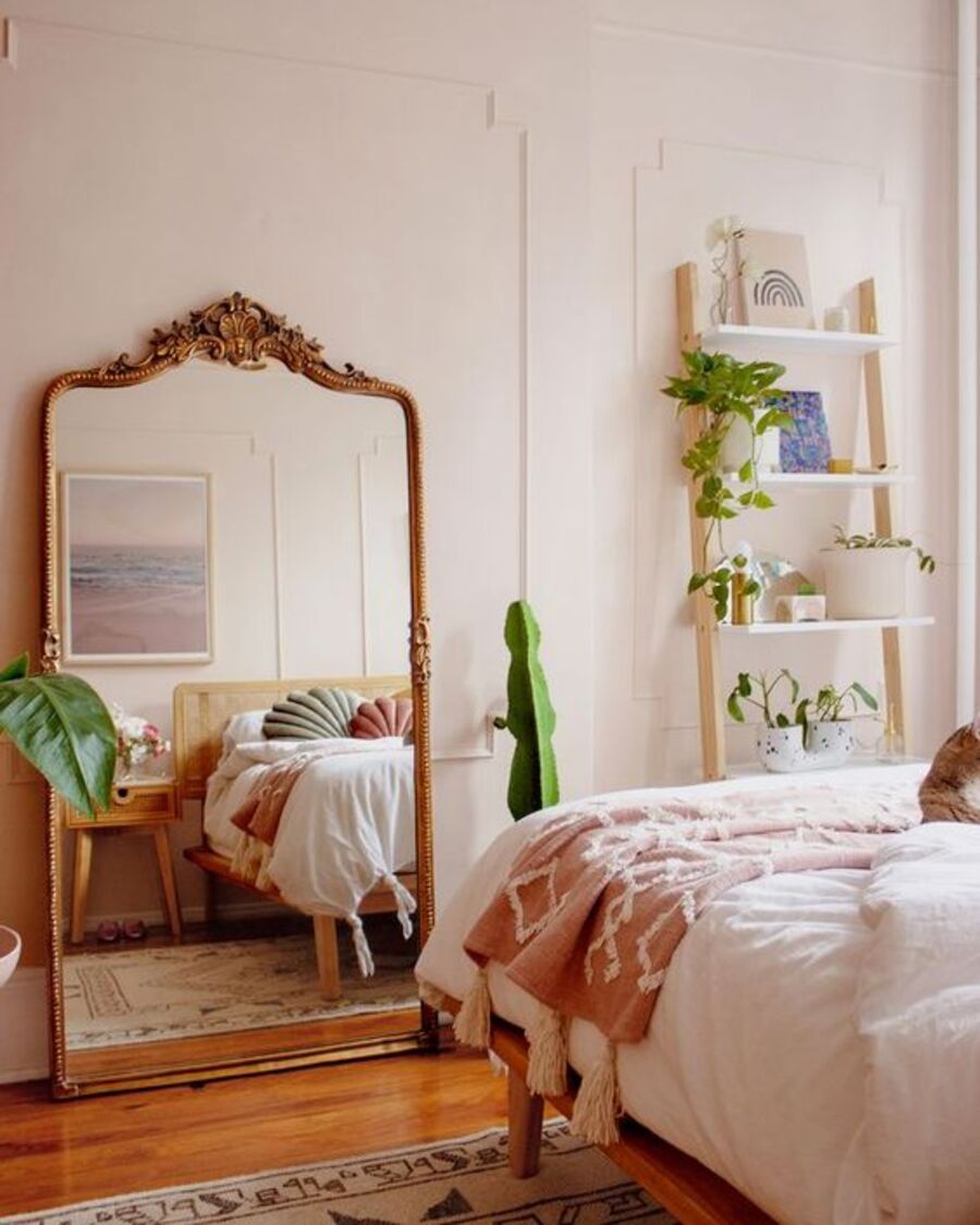 Dormitorio de estilo vintage con un gran espejo con marco dorado y detalles clásicos. Está apoyado sobre el suelo de madera y refleja una cama con plumón blanco, manta rosada y cojines rosado y verde claro. Junto al espejo se asoman dos plantas grandes.
