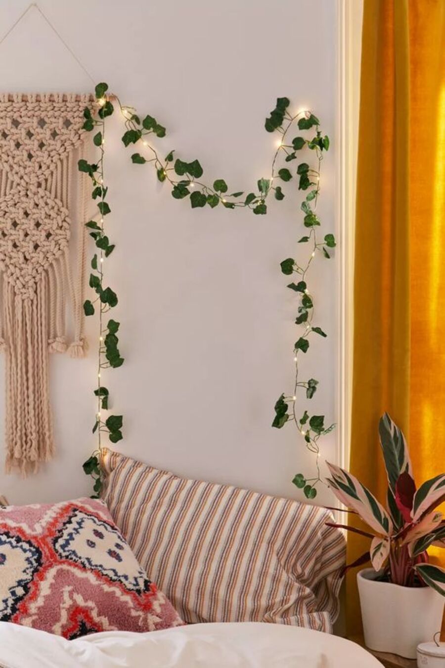 Detalle de un rincón boho chic donde se aprecia parte de una cortina mostaza, cojines estampados de colores, una planta, un adorno de pared de macramé y una planta con luces decorativas.
