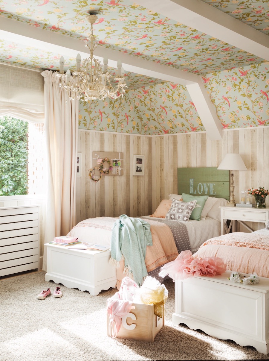 Dormitorio infantil con dos camas con mantas y cojines de colores pastel. Es suelo es alfombrado y el techo, que tiene vigas blancas, también está forrado en papel mural celeste con flores y hojas verdes, rosadas y amarillas. 
