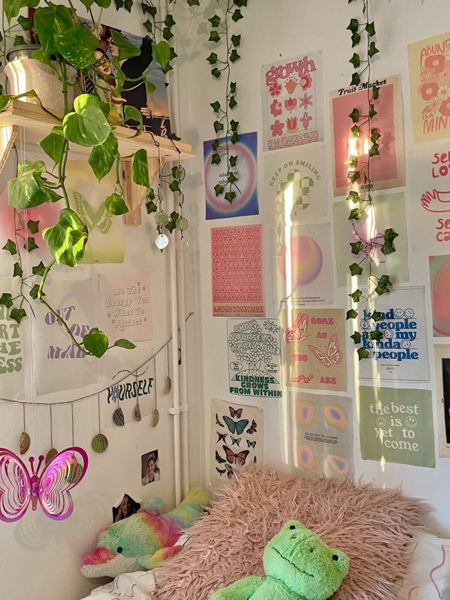 Dormitorio adolescente con pared blanca y posters de colores pasteles, desde rosadas hasta verdes. También cuelgan plantas y sobre la cama hay un cojín rosado peludo, un peluche de rana verde y un peluche de delfín multicolor.