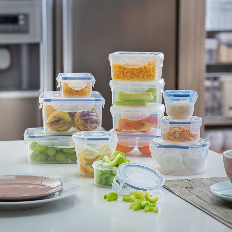 12 contenedores plásticos, herméticos y transparentes con distintos alimentos en su interior, tales como: frutas, verduras, huevos y cereales. Está sobre un mesón de una cocina.