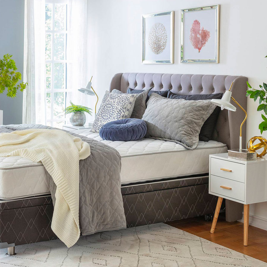 Dormitorio con cama y respaldo de tela estilo capitoné gris. Sobre la cama hay una frazada gris, una manta blanca y cojines y almohadas en tonos neutros.