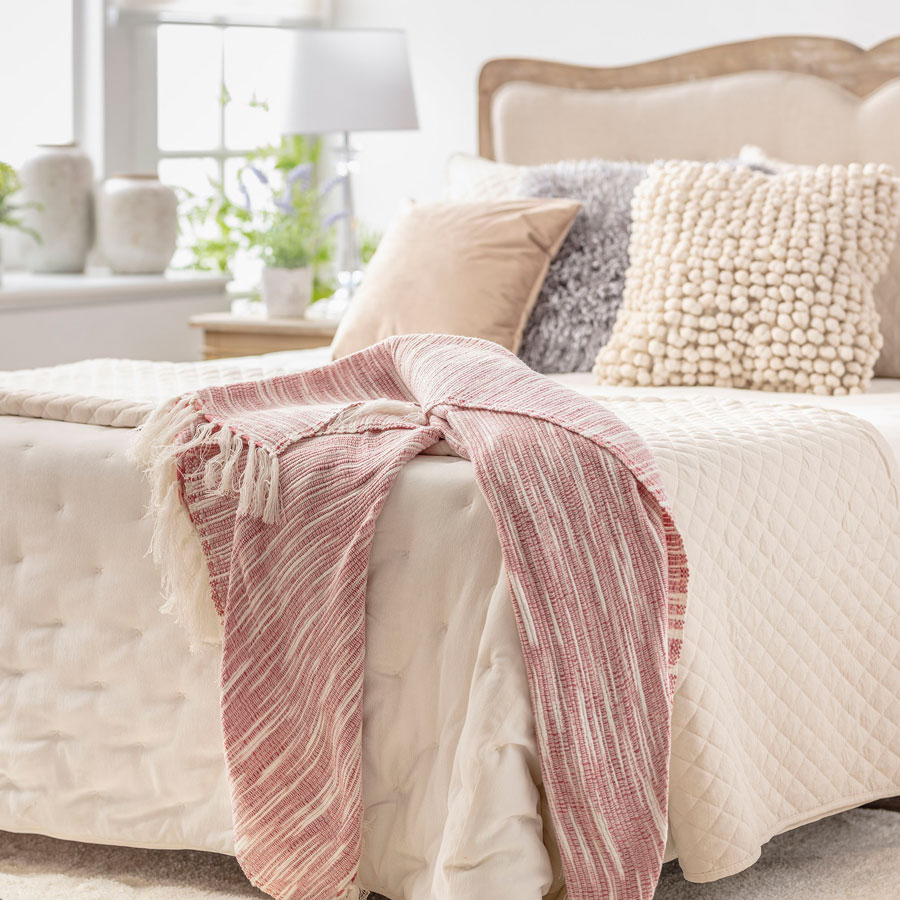 Detalle de una cama con cobertor color crema, cojines en tonos neutros y una manta rayada blanca con rosado. En el piso hay una alfombra peluda blanca.