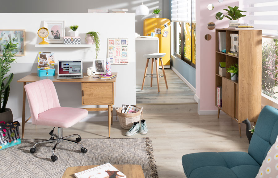 Zona de trabajo con un escritorio y estante de madera, silla de terciopelo rosado con ruedas. El suelo es piso flotante y sobre él hay una alfombra gris con detalles beige. Hacia el fondo se ve parte de una cocina, también con piso flotante. 