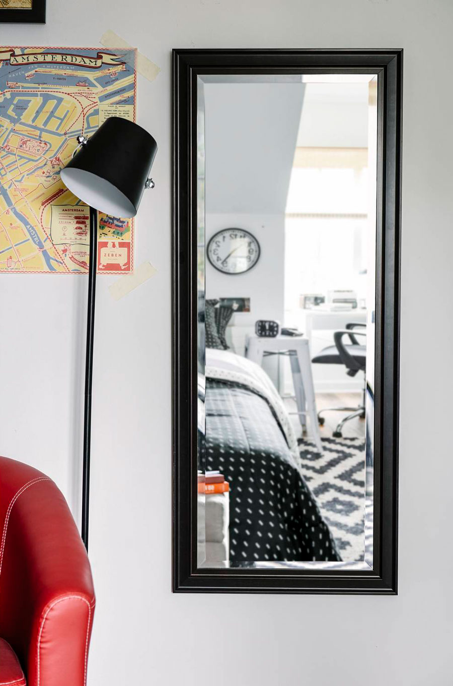 Detalle de una pared de un dormitorio con un espejo rectangular de cuerpo completo y marco negro. Al costado hay una lámpara de pie negra, un mapa pegado en la pared y parte de un sofá rojo. En el reflejo del espejo se ve parte de la cama y escritorio.