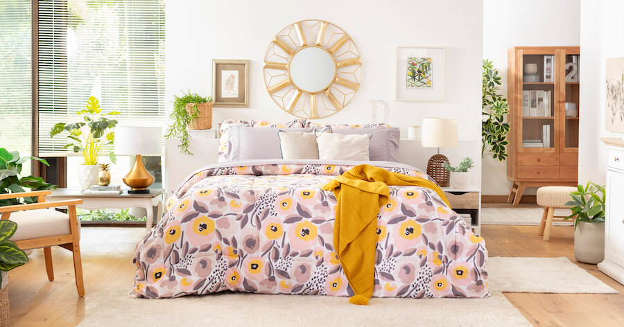 Dormitorio de paredes blancas, piso de madera y cama con un plumón de flores amarillas con hojas moradas. En la cabecera hay un espejo redondo con un marco con diseño dorado. 
