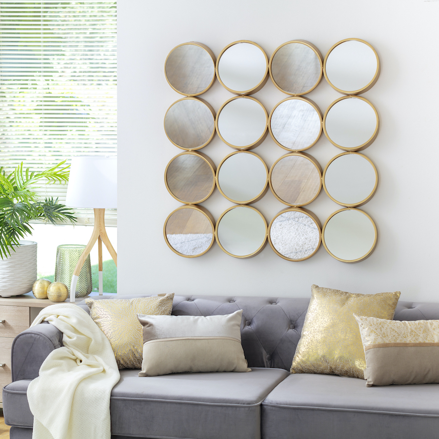 Living con sofá gris, cojines dorados y manta blanca. En el muro sobre el sofá hay una serie de pequeños espejos redondos con borde dorado, unidos entre sí formando una especie de cuadrado, ideales para el Feng Shui.