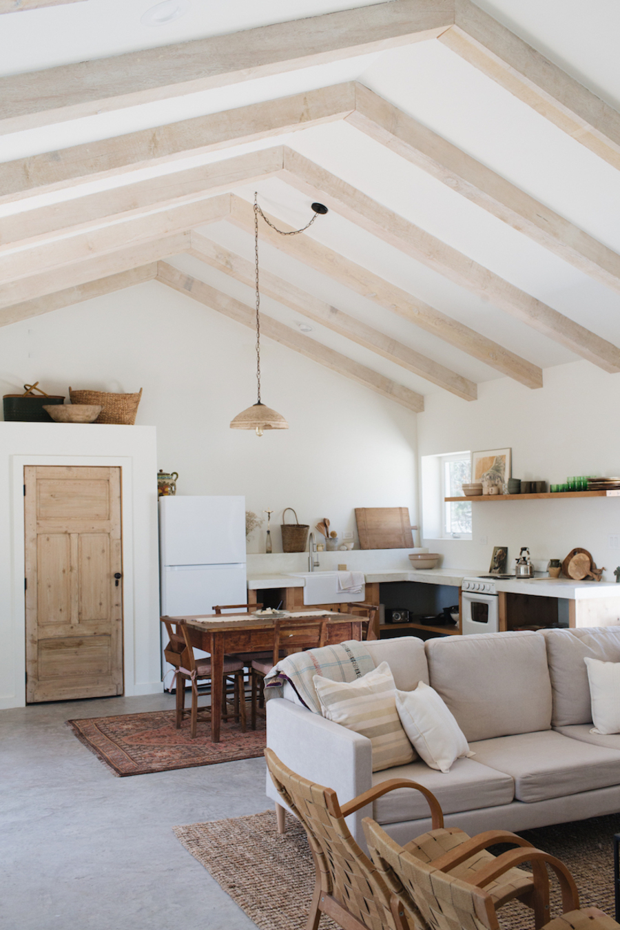 Cocina de concepto abierto con un comedor de madera y living contiguo. En el techo, que es blanco, hay una serie de vigas de madera clara. 