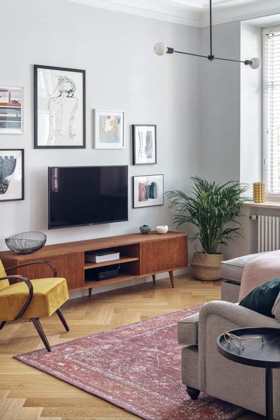Sala de estar con paredes gris claro, piso de madera y rack de madera oscura. Sobre él hay un muro galería y una televisión que cuelga sobre el rack. Frente a ella hay un sofá beige. 