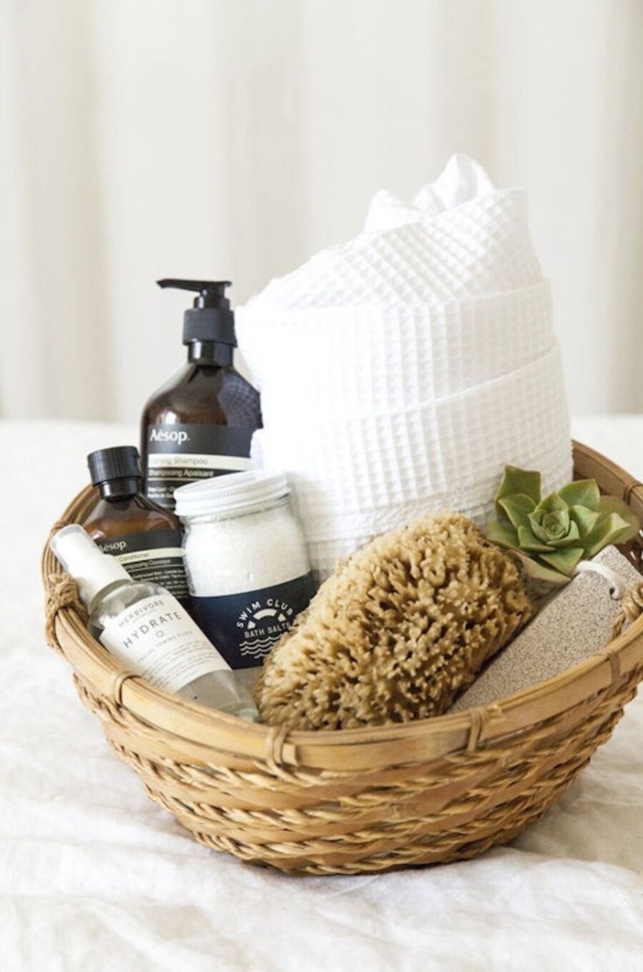 Detalle de una cesta de fibras naturales con diversos artículos de higiene en su interior, desde una toalla y esponja hasta shampoo y otros cosméticos.