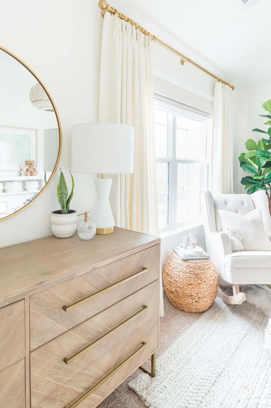 Espacio del living con una poltrona y alfombra blancas, cajonera de madera y una ventana con cortinas de tela blanca colgadas en una barra dorada.