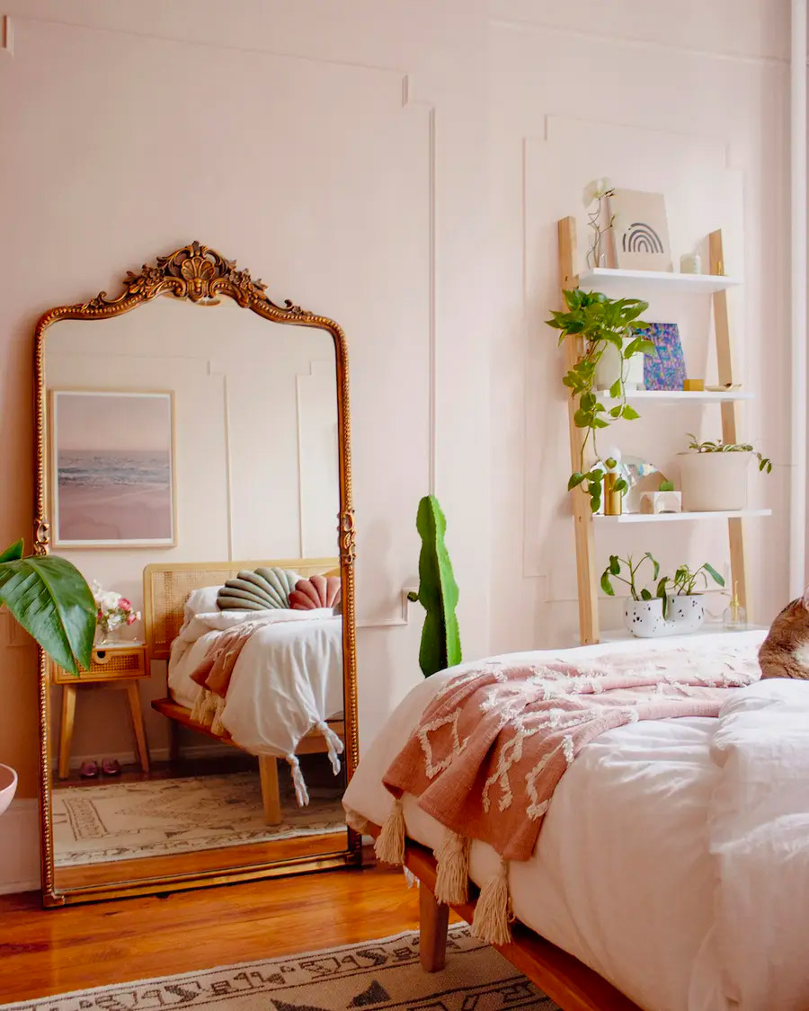 Dormitorio con muros blancos y piso de madera. La ropa de cama es blanca y terracota y frente a ella hay un gran espejo de estilo clásico que refleja parte de la cama.