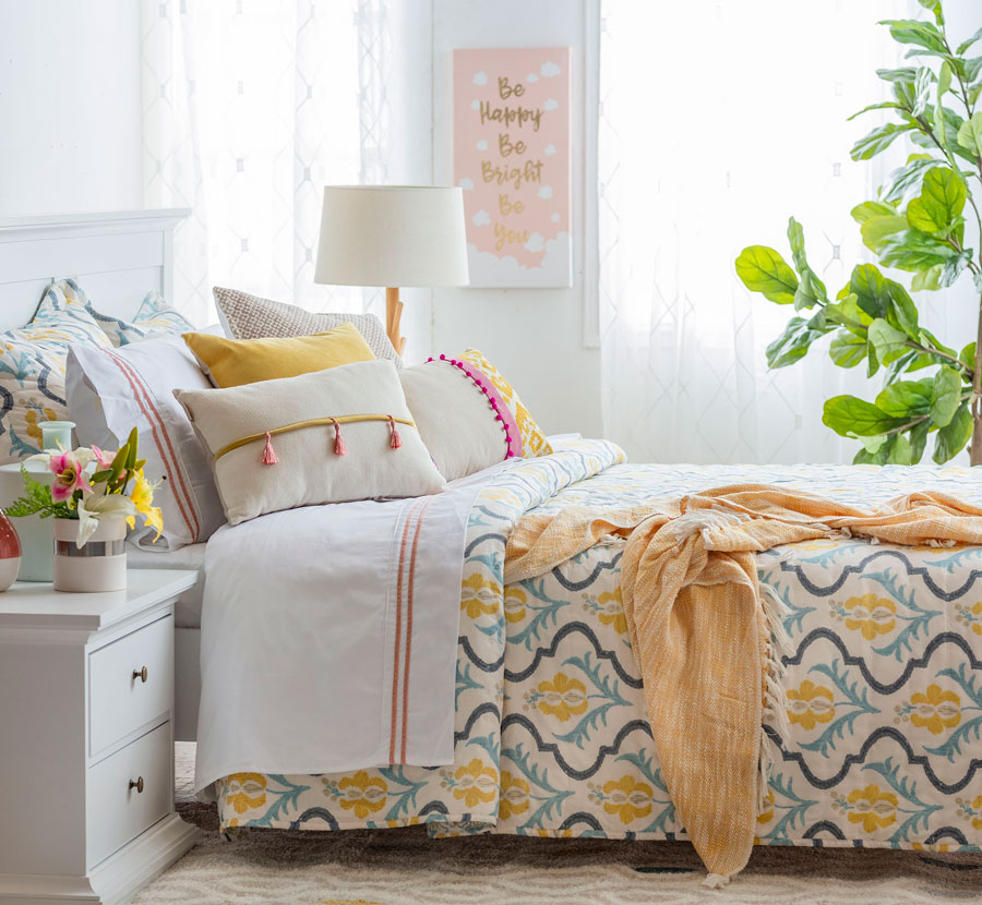 Dormitorio con una cama con cojines de colores y cobertor con estampado con diseño azul, celeste, amarillo y beige. Hacia el fondo hay dos ventanas con visillos blancos que filtran la entrada de la luz.