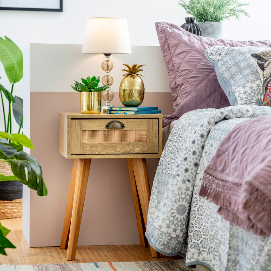 Detalle de un velador de madera con decoración dorada sobre él. Está junto a una cama con cojines y cobertor en tonos azulados y lila. 