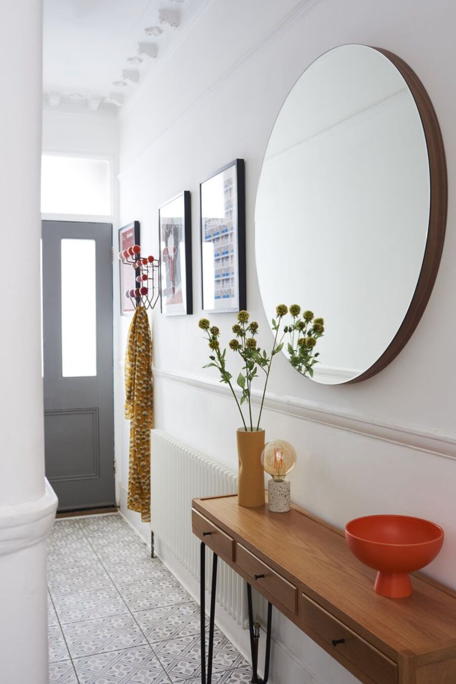 Pasillo de muros blancos con un gran espejo redondo en una de sus paredes, debajo hay un arrimo de madera con patas metálicas negras. Al costado del espejo hay tres cuadros con ilustraciones coloridas y un perchero rojo.