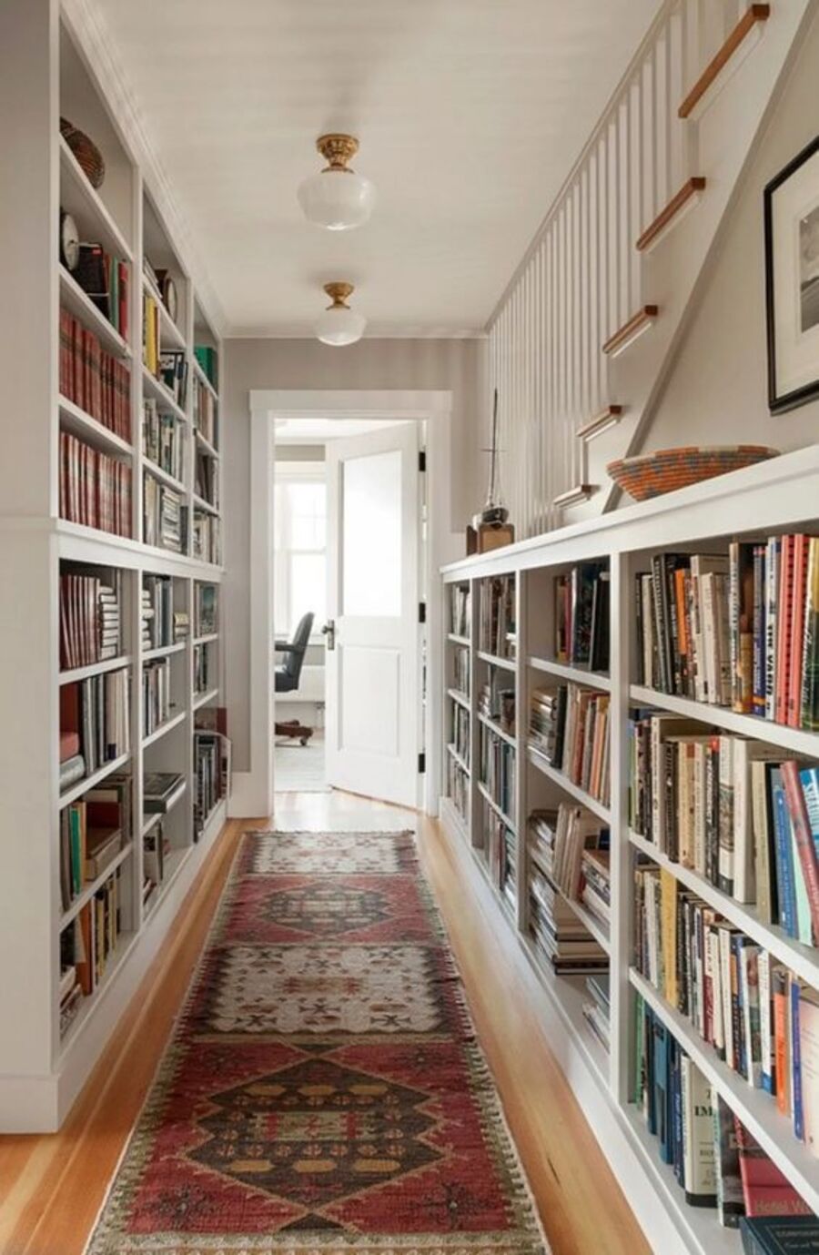Pasillo con piso de madera con alfombra estampada en tonos rojizos. En los muros hay estantes blancos con libros de colores. 