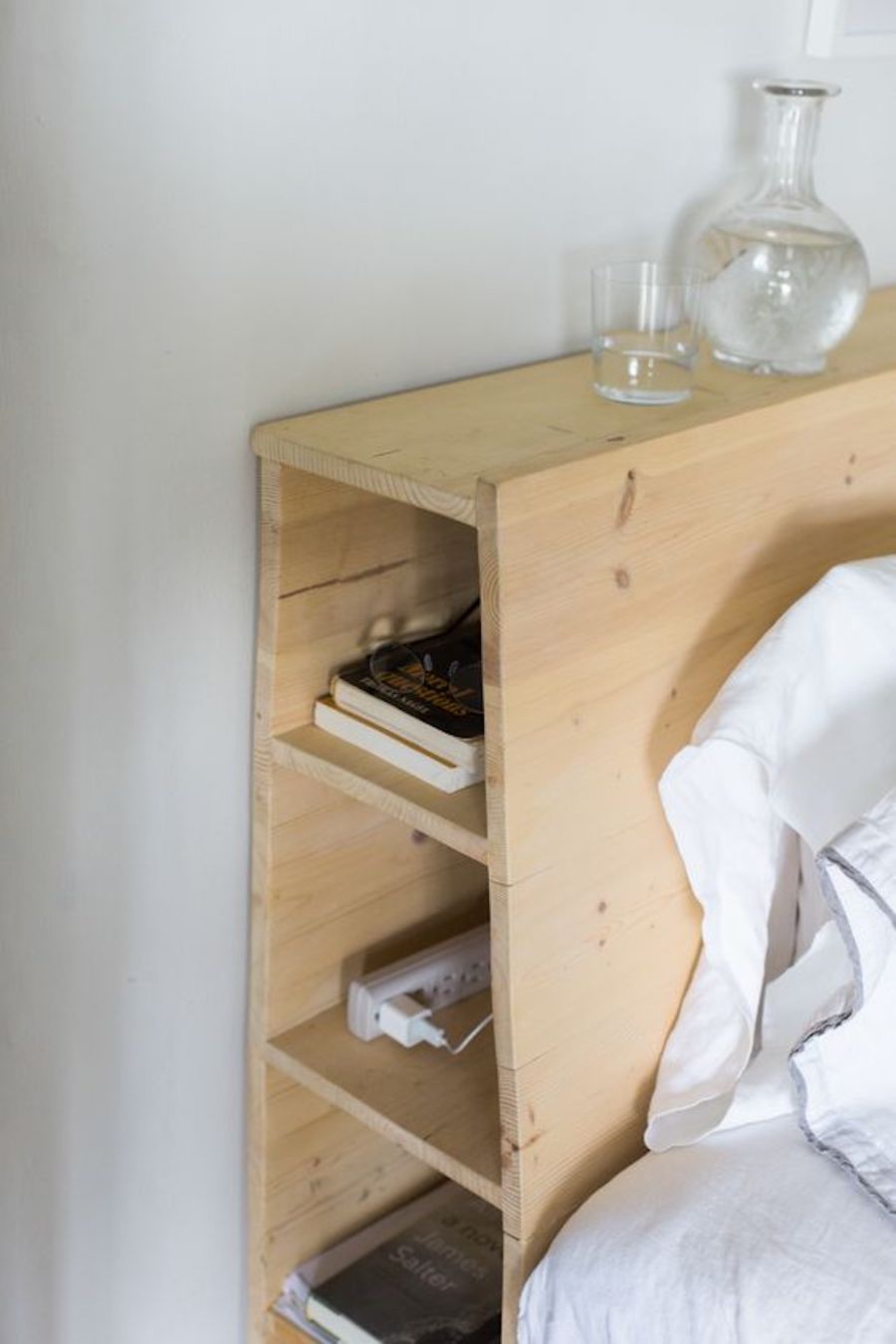 Detalle de un respaldo de cama de madera clara, en cuyo costado hay espacio para poner libros. Sobre él hay un vaso y jarro con agua. La sábana y almohada de la cama son blancas.