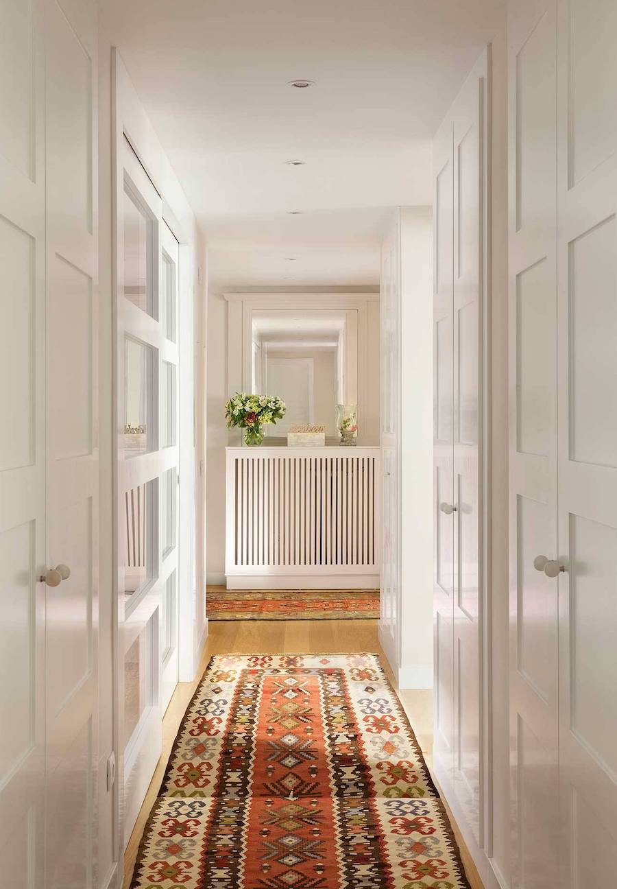 Pasillo con paredes y techos blancos y piso de madera con una larga alfombra estampado en colores rojizos. 